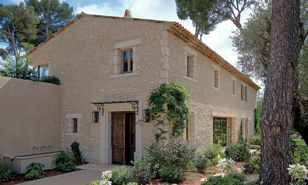 Casa in pietra in stile rustico immersa nel verde, con finestre incorniciate in pietra chiara, porta in legno scuro e alberi alti che circondano il giardino.